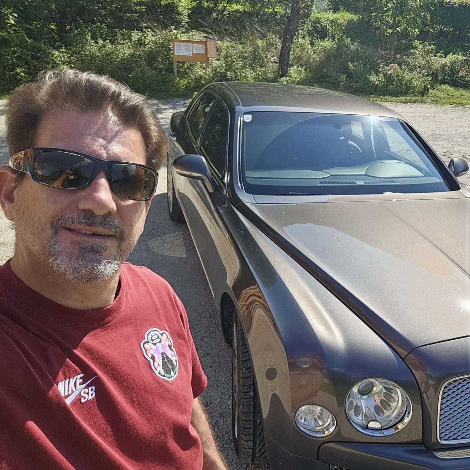 Man in sunglasses and red shirt standing next to a Bentley car outdoors.